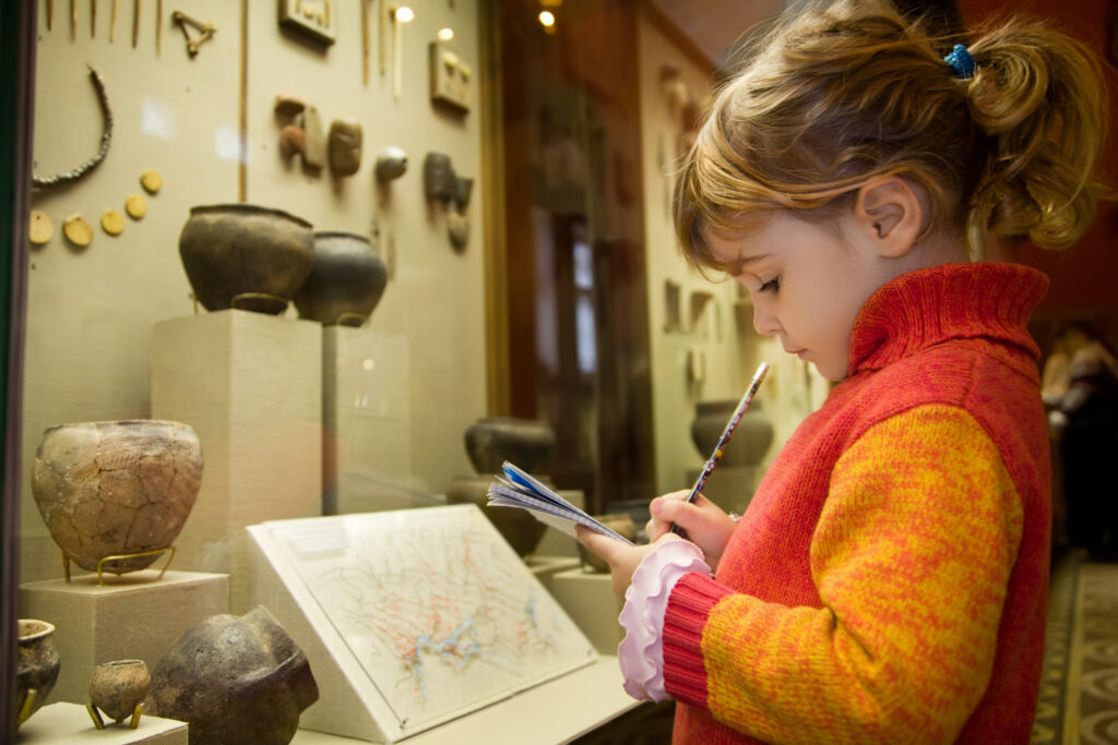 little girl writes to writing-books at excursion in historical museum near exhibits of ancient relics in glass cases