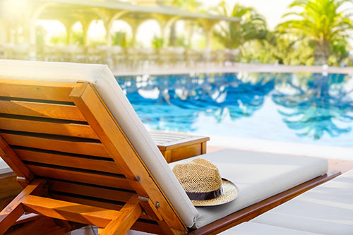 Beach lounger with straw hat and swimming pool with reflection of palm trees at the resort.