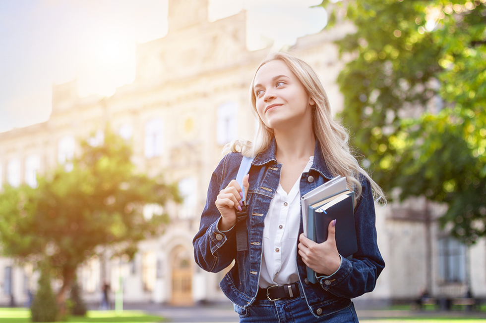 Smiling young female student is holding educational books, standing against background of college building at campus. Happy blond girl near university. High school lifestyle. Education concept.
